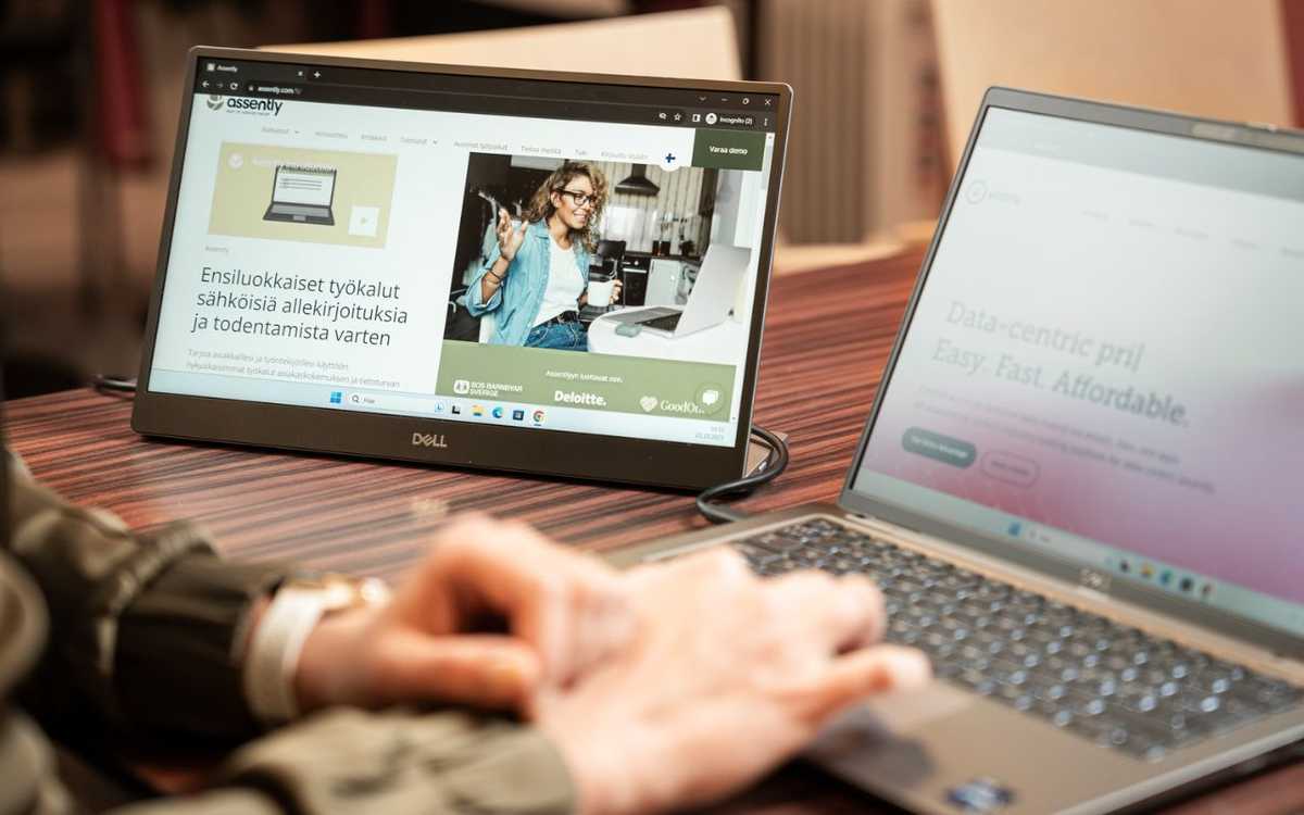 Henkilö käyttää kannettavaa tietokonetta. Tietokoneeseen on yhdistetty ulkoinen näyttö, jolla näkyy sähköisten allekirjoitusten palvelun Assentlyn kotisivut.
